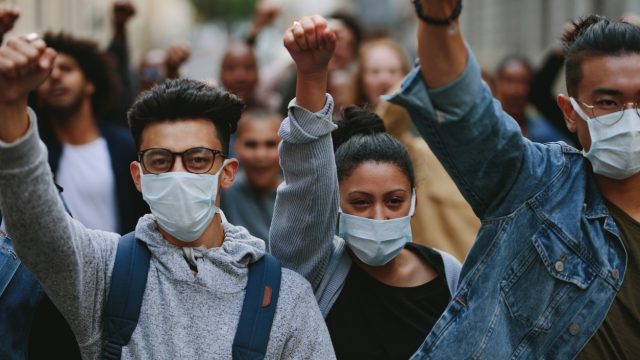 A group of ethnically diverse young people raise their hands at a protest while wearing face masks