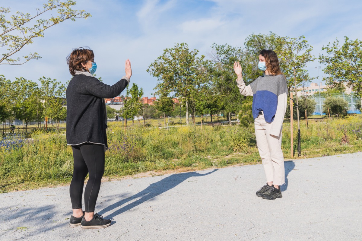 two white women in face masks waving at each other from a distance in a park