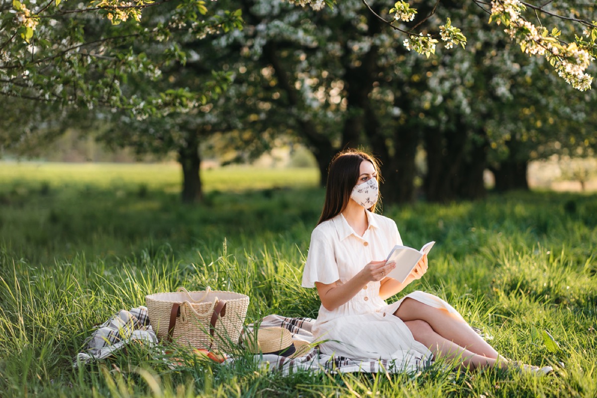 white woman with a face mask reading a book next to a picnic basket on a blanket