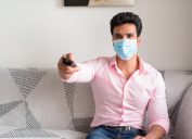 man watching tv and holding remote while wearing a mask