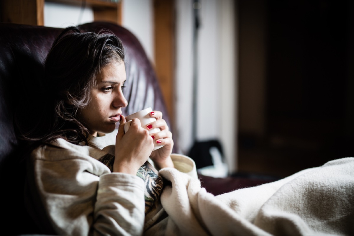 sick white woman drinking tea under a blanket