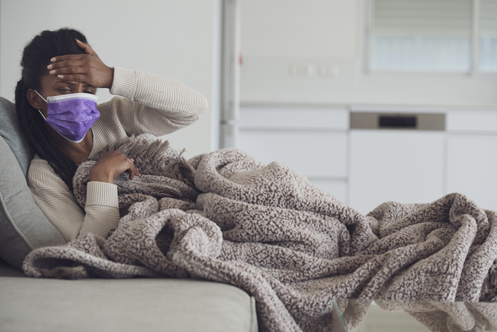 A young woman wearing purple face mask lying on the couch under a blanket and checking her forehead for a fever