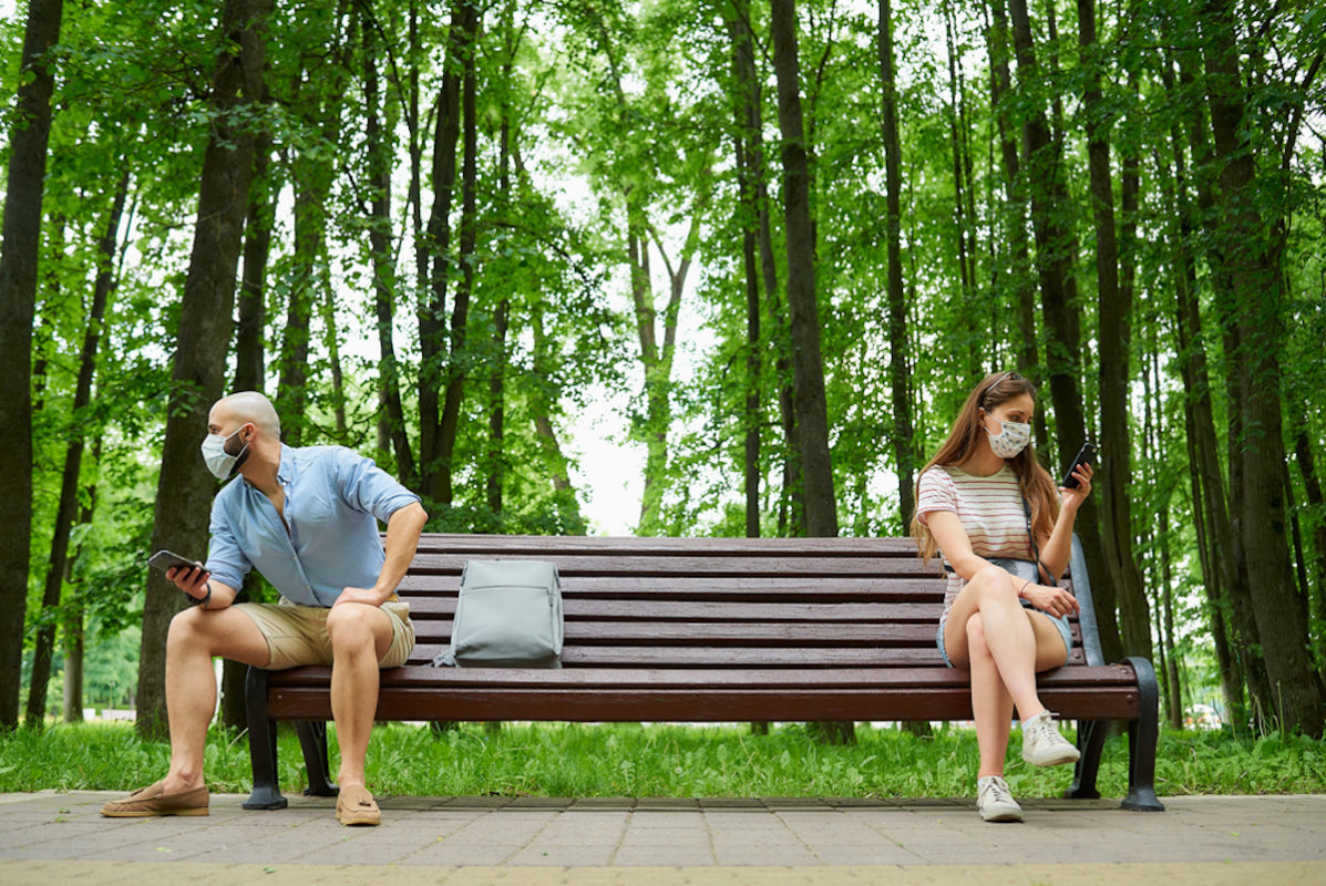 two people sitting on opposite ends of bench in park with face masks on