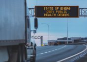 State of Emergency Obey Public Health Orders on an overhead highway sign during the Coronavirus pandemic, out of focus semi truck in foreground.