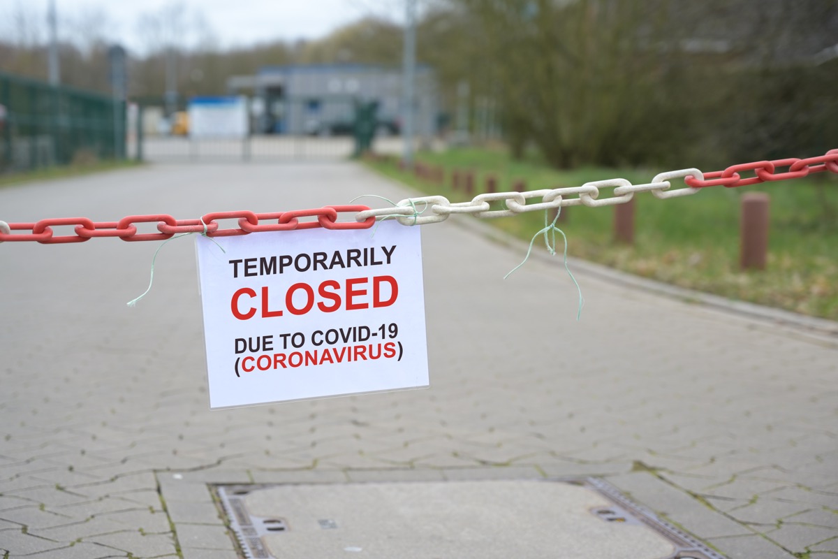 Red white chain barrier and sign with text Temporarily Closed due to Covid-19 Coronavirus, in front of a blurred company, countrywide pandemic lock down, copy space, selected focus