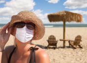 A woman in sunglasses and a hat wearing a face mask on a sunny beach
