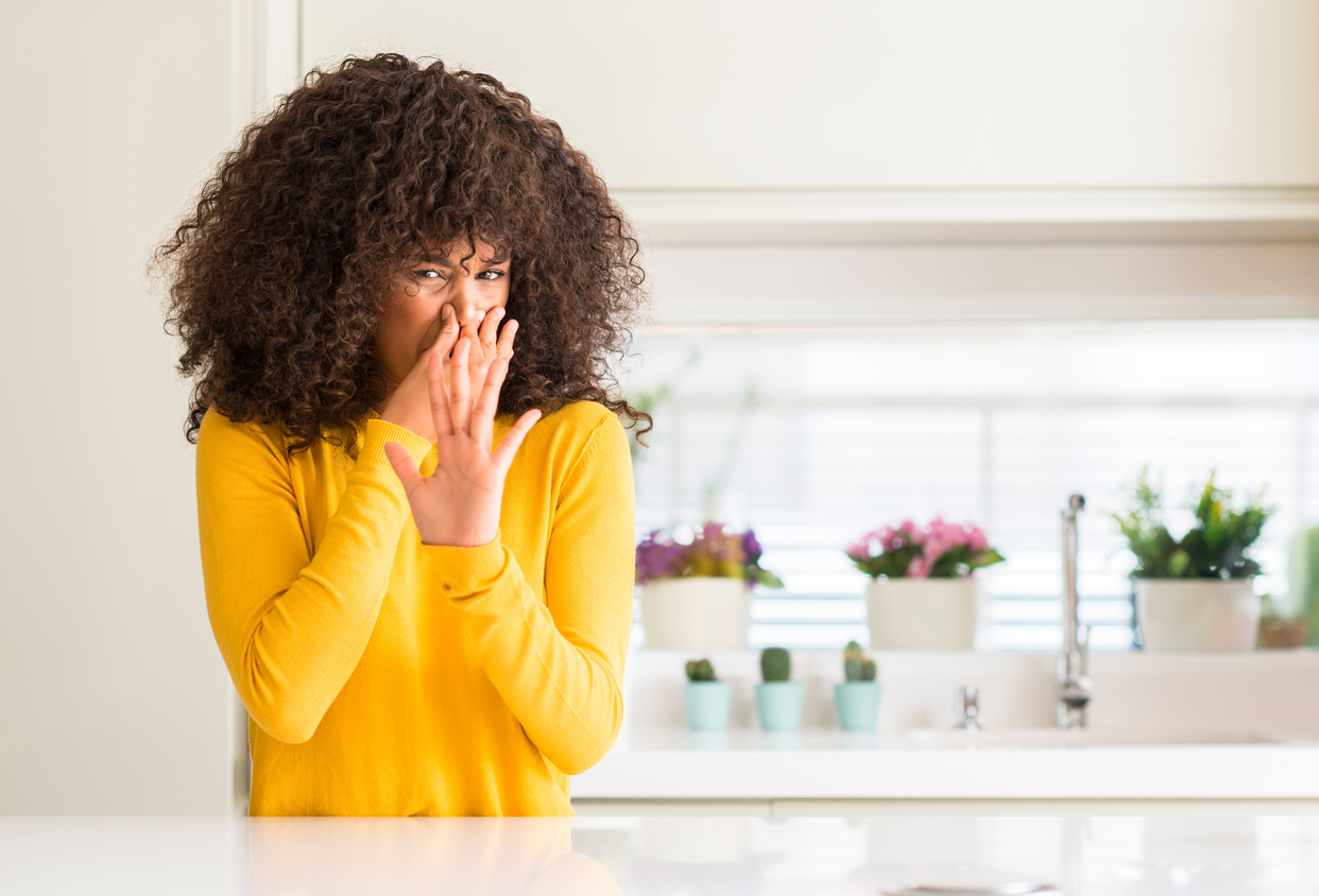 African american woman wearing yellow sweater at kitchen smelling something stinky and holding her nose