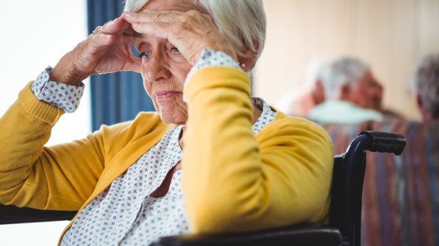 A senior woman sitting in a wheelchair holding her head looking confused and worried