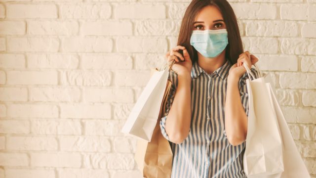 Happy girl shopper in protective face mask with paper bags in hands. Young woman in medical face mask holding shopping bags. Girl shopaholic in mall. Sale, discount, coronavirus COVID-19, copy space