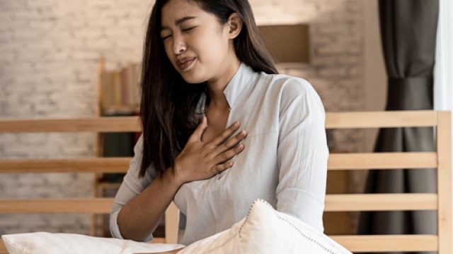 An Asian woman sitting in bed with her hand on her chest showing difficulty breathing