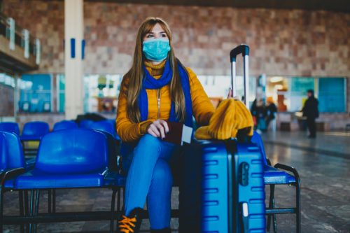 A woman wearing a face mask sits next to her blue suitcase in a travel lounge