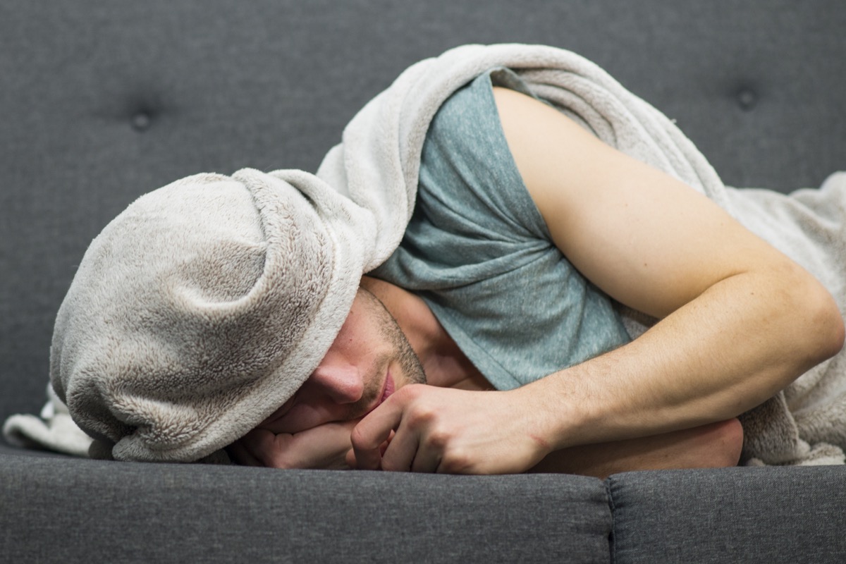 young man wrapping a blanket around his head feeling depressed as he lays sadly on a grey couch alone.
