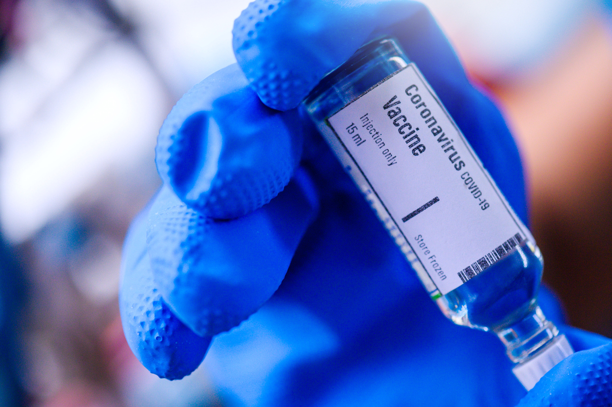 A close up of a blue gloved hand holding a glass vile of coronavirus vaccine dosage