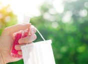 closeup of woman's hand outside holding pink bottle of hand sanitizer and mask in hand