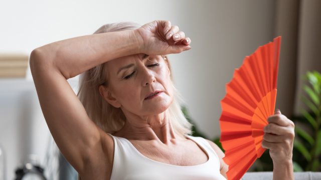 overheated senior woman waving fan due to heat while sitting on couch