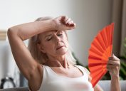 overheated senior woman waving fan due to heat while sitting on couch