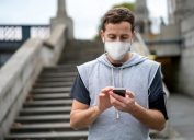 Portrait of a male jogger outdoors wearing a facemask and listening to music while working out in quarantine – COVID-19 lifestyle concepts