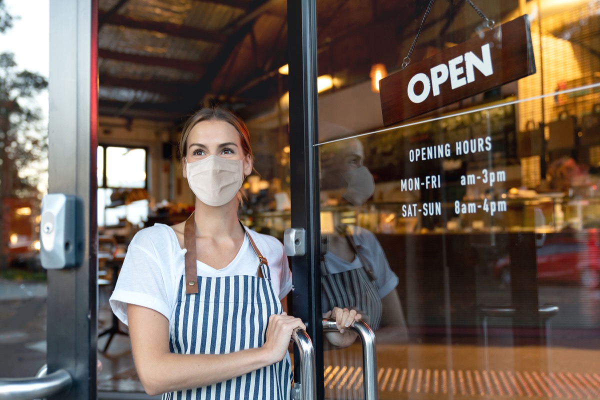 Happy business owner opening the door at a cafe wearing a facemask to avoid the spread of coronavirus – reopening after COVID-19 concepts