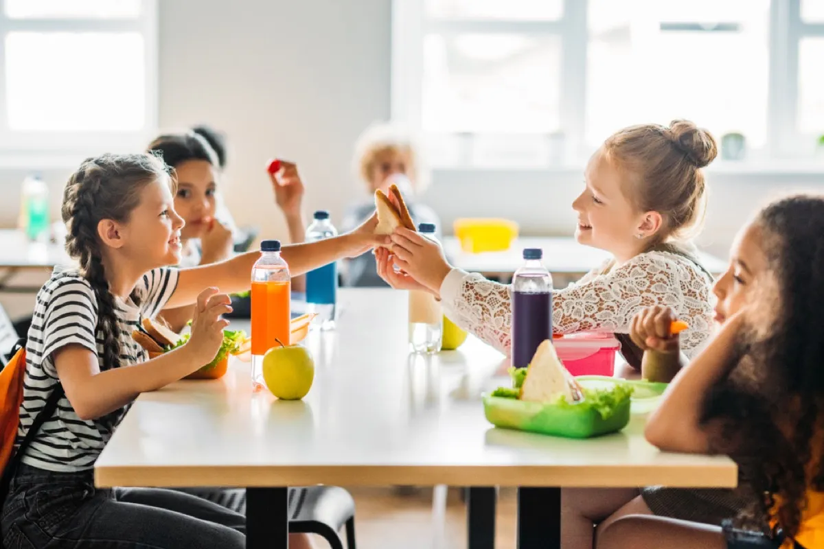 two girls sharing a sandwich and sitting with friends at a school cafeteria