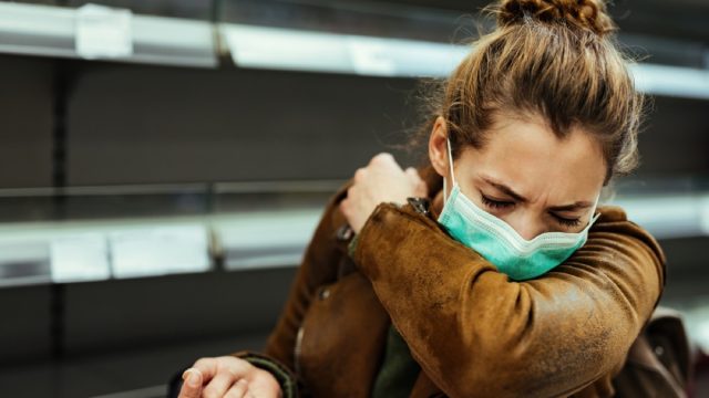 woman with mask coughing into her arm in front of empty shelf at supermarket