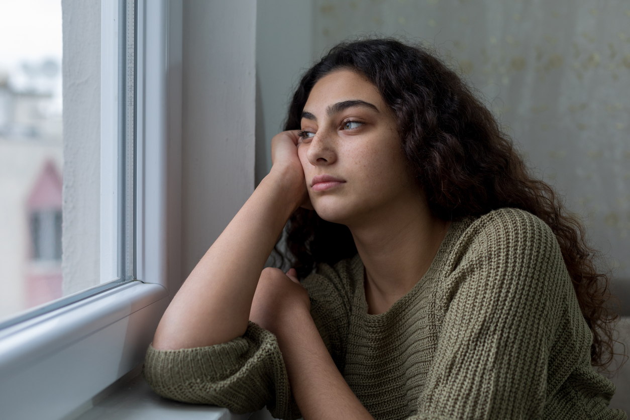 A teenage girl with dark hair looks out a window with a sad look on her face.