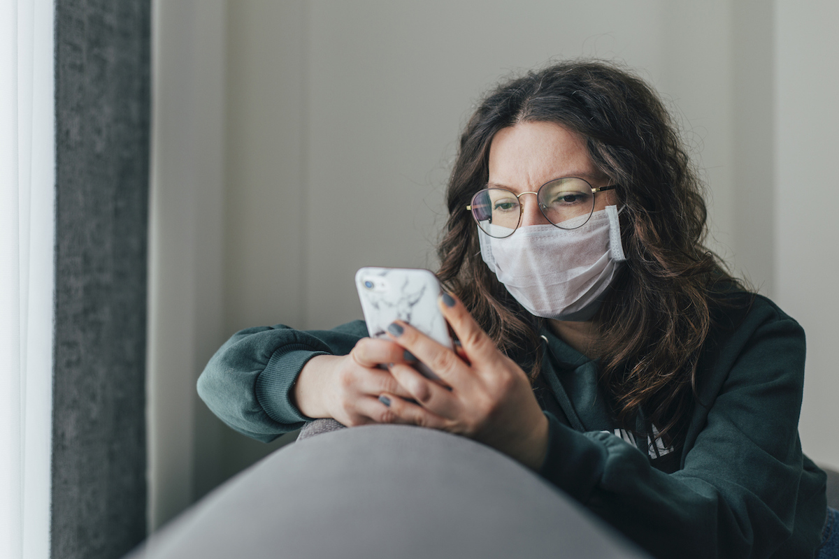 Young woman covering her face with surgical mask and using smartphone to find information about coronavirus spread on internet