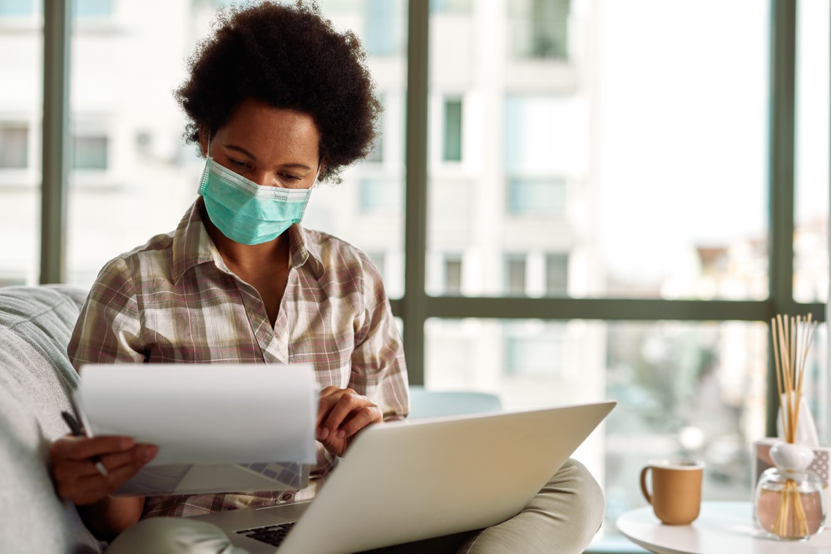 woman with face mask reading business reports while working on laptop at home during virus pandemic.