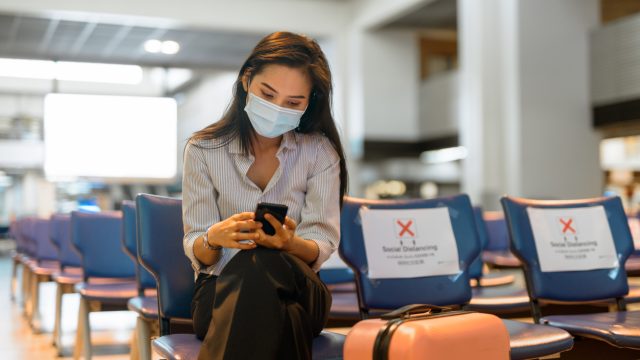 A young woman sitting in an airport while wearing a face mask, sitting next to socially distanced seats