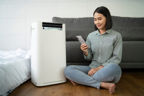 A young woman sits next to an air purifier in her apartment while looking at her phone