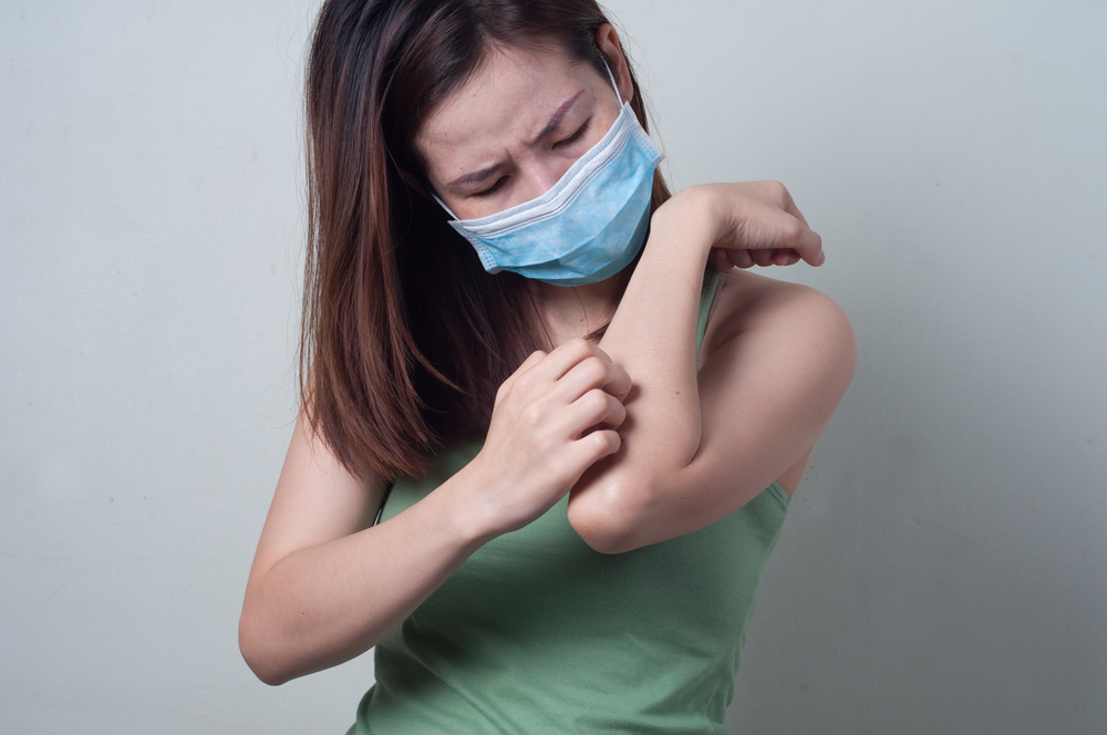 young asian woman scratching arm while wearing surgical mask