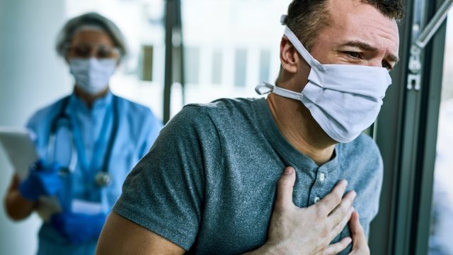 Male patient wearing face mask and feeling chest pain while being at the hospital during coronavirus epidemic. Healthcare worker is in the background.