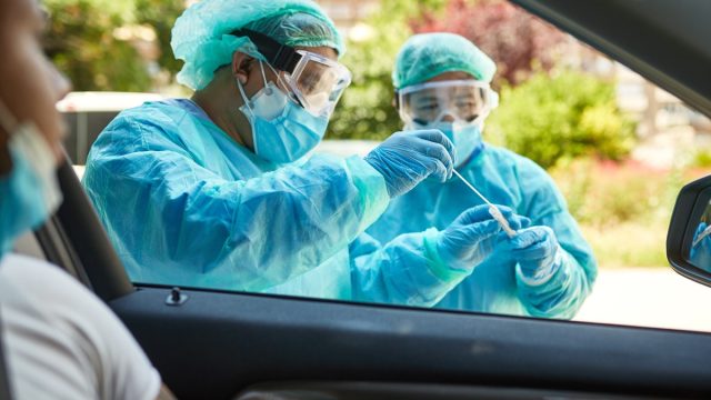 Young man sitting in car watching as hospital team in protective workwear place nasal swab specimen in sterile container.
