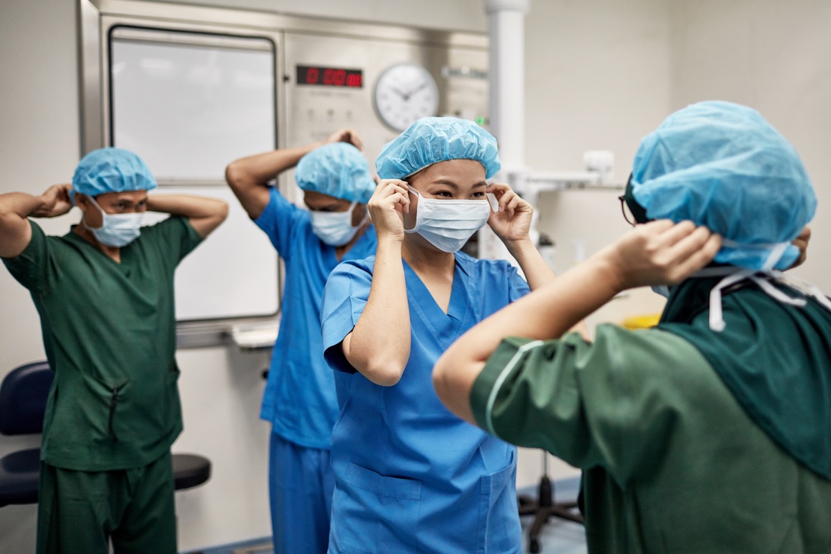 medical team in 30s and 40s dressed in blue and green medical scrubs, surgical caps, and smiling as they put on surgical masks in operating room.