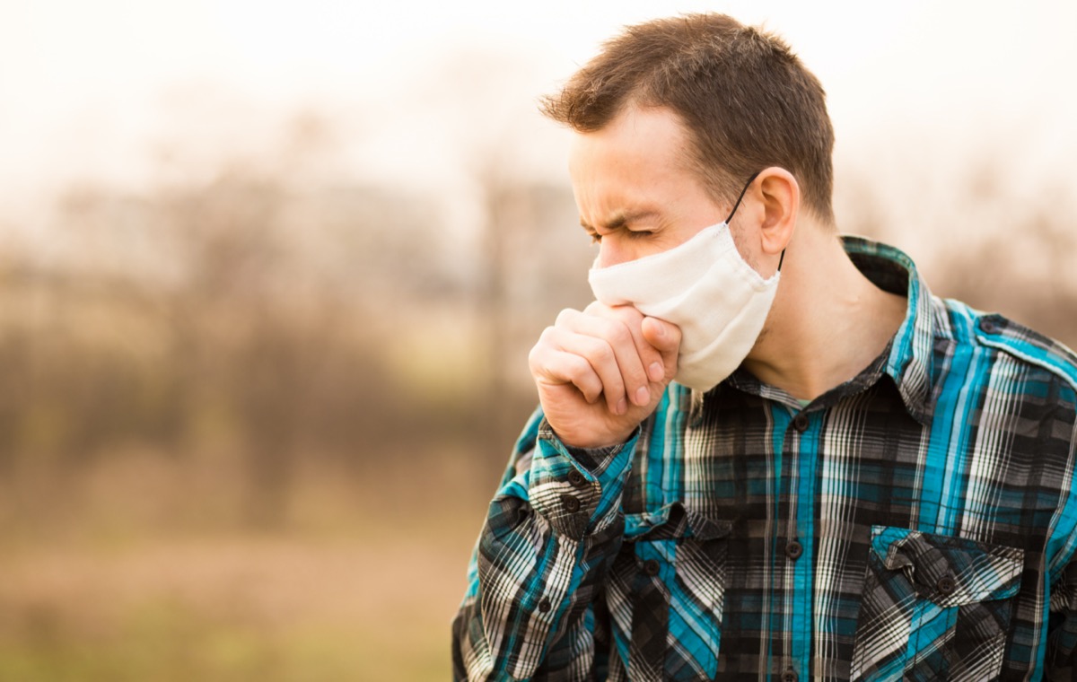 young white man standing outdoors wearing face mask and coughing
