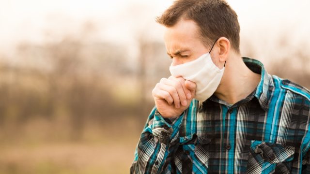 young man standing outdoors wearing face mask and coughing