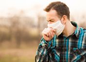 young man standing outdoors wearing face mask and coughing