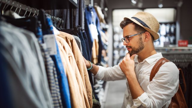A young man in a hat and glasses shops for clothes in a store