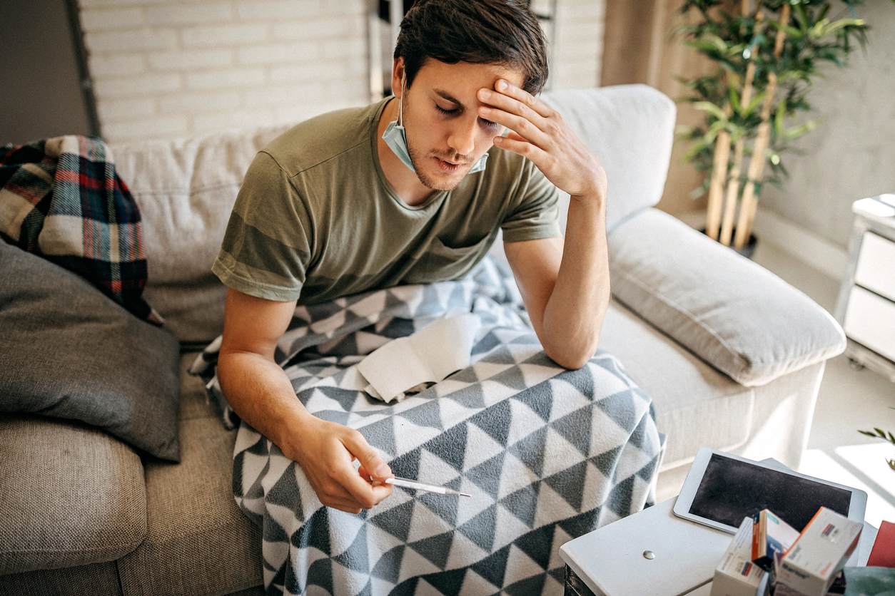 A young man with coronavirus symptoms sitting on the couch reading a thermometer with a face mask pulled down under his chin