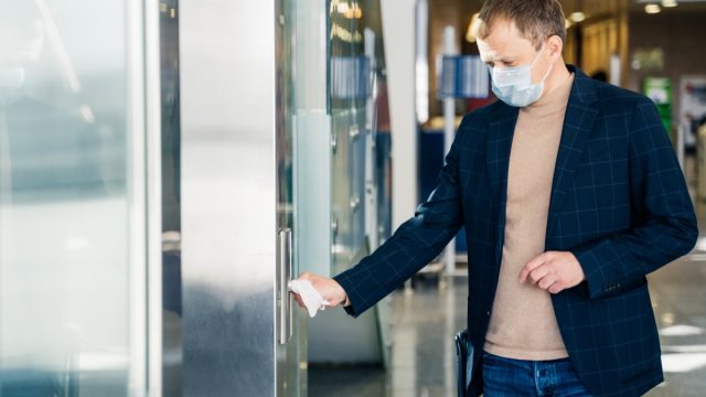 Man protects hands from direct contact with door handle, uses paper napkin from conntracting virus through contact with surface, wears disposable medical mask to prevent covid19, poses in public place