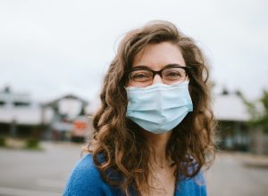 A young woman smiles while wearing a protective medical mask outdoors. Happy still in the face of the global pandemic of Covid-19.