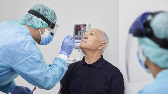 A older man in a blue short has his nose swabbed by a health care worker in full protective gear for a coronavirus test