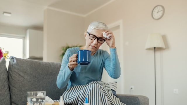 An older woman sits on the couch with symptoms of the common cold, holding a mug and tissue