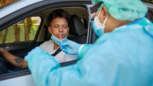 A health care worker in full protective gear performs a nasal swab on a young man sitting in his car to test for coronavirus