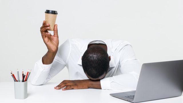 Black man with head on desk holding up coffee