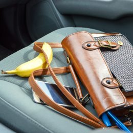 This is an image of a brown purse on the passenger seat of a car with contents spilling out. There is a pen, makeup item, phone and banana laid out around the purse. The car interior is grey.
