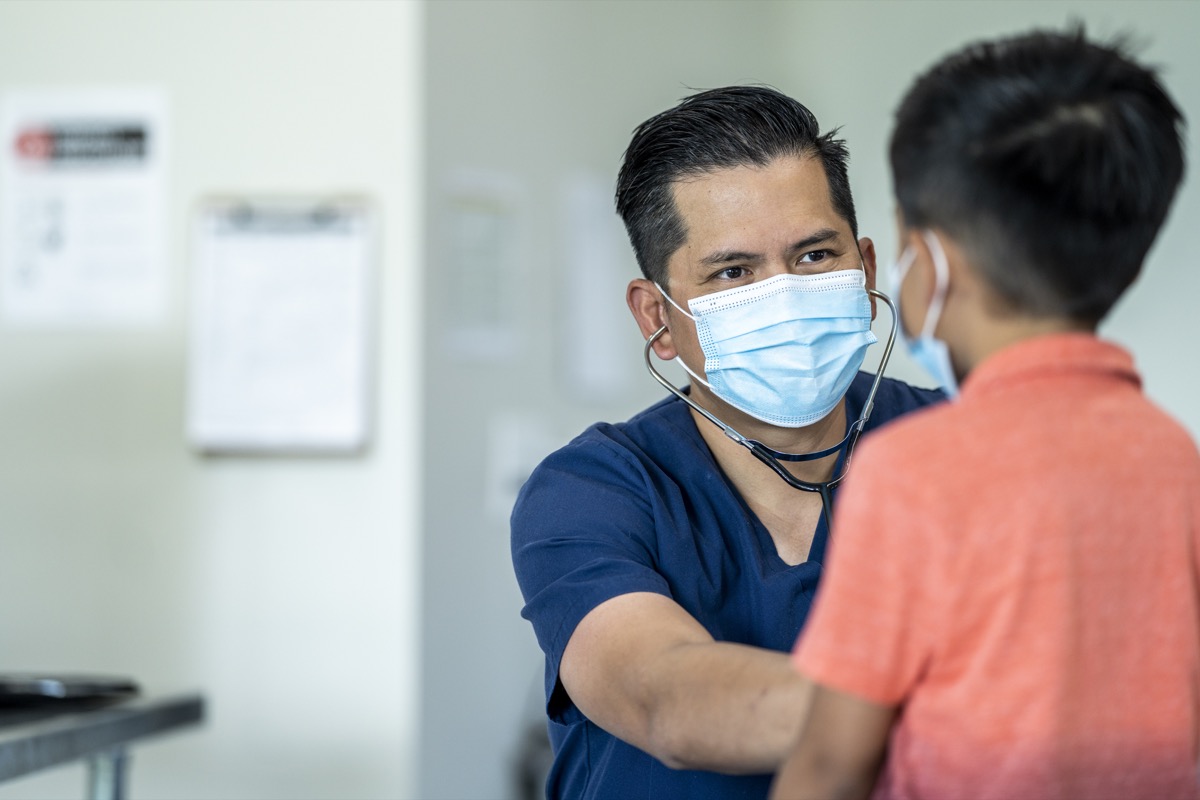 Male doctor wearing a mask and gloves during a checkup with a young, 8 year old boy because of the COVID-19 outbreak.