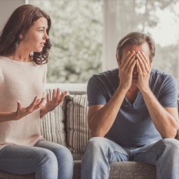 Couple of adults are quarreling while sitting on couch at home, man is covering his face