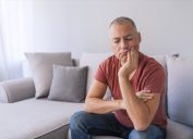 Photo of Depressed ill man having toothache and touching cheek. Mature man suffering from tooth pain, caries. Handsome gray hair male suffering from toothache, closeup. Portrait of casual 46s mature man toothache with painful expression, sitting on sofa at home,