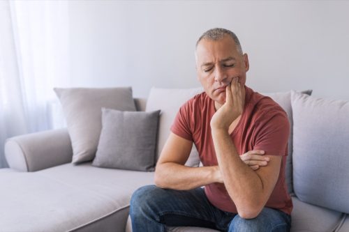 Photo of Depressed ill man having toothache and touching cheek. Mature man suffering from tooth pain, caries. Handsome gray hair male suffering from toothache, closeup. Portrait of casual 46s mature man toothache with painful expression, sitting on sofa at home,