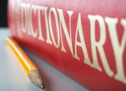 A red dictionary sitting on a table top with a pencil next to it.